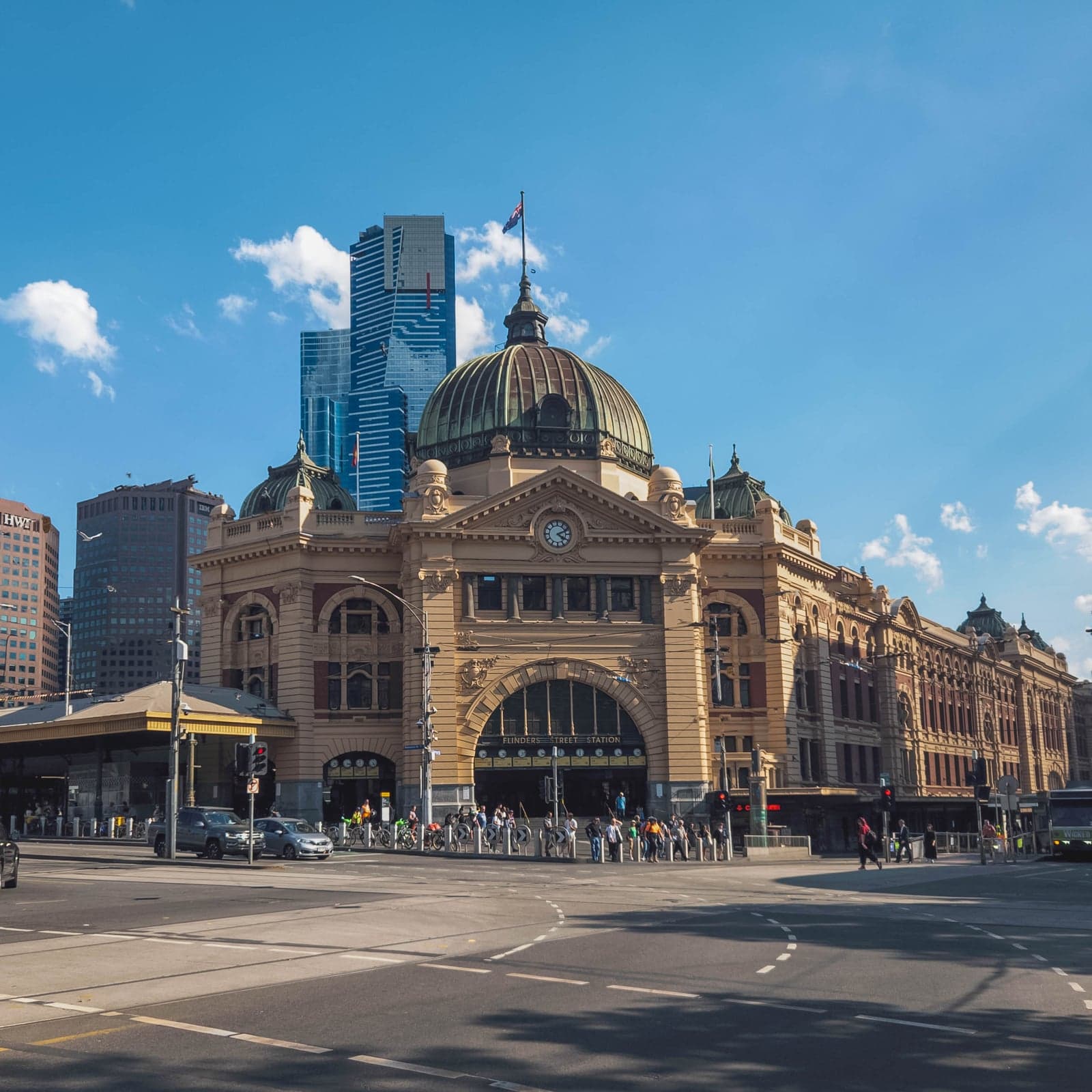 Flinders Street Station in Melbourne — iconic yellow-ochre sandstone facade with central clock, tripartite arched entrance and copper-domed clocktower, with Eureka Tower and Melbourne's city skyline behind.
